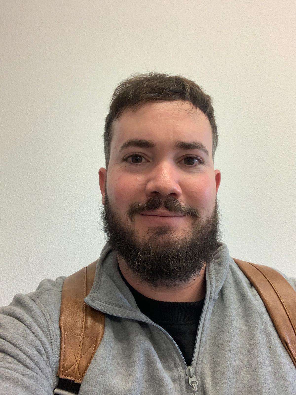 Hombre sonriente con barba y mochila frente a pared blanca